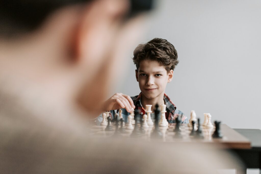 In this heartwarming snapshot, a young child is immersed in the fascinating world of chess, seated at a chessboard with unwavering focus. The child, a budding chess enthusiast, gazes directly at the camera with an infectious and confident smile. Their eyes sparkle with the joy of the game, and their expression radiates a sense of accomplishment and pride.